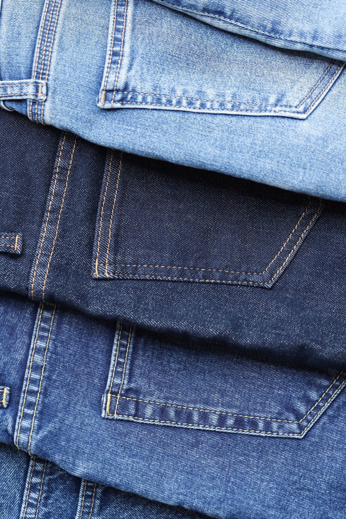 Close-up of folded denim jeans showing three different blue washes: a dark indigo, a medium blue, and a light wash, with visible stitching and pocket details.
